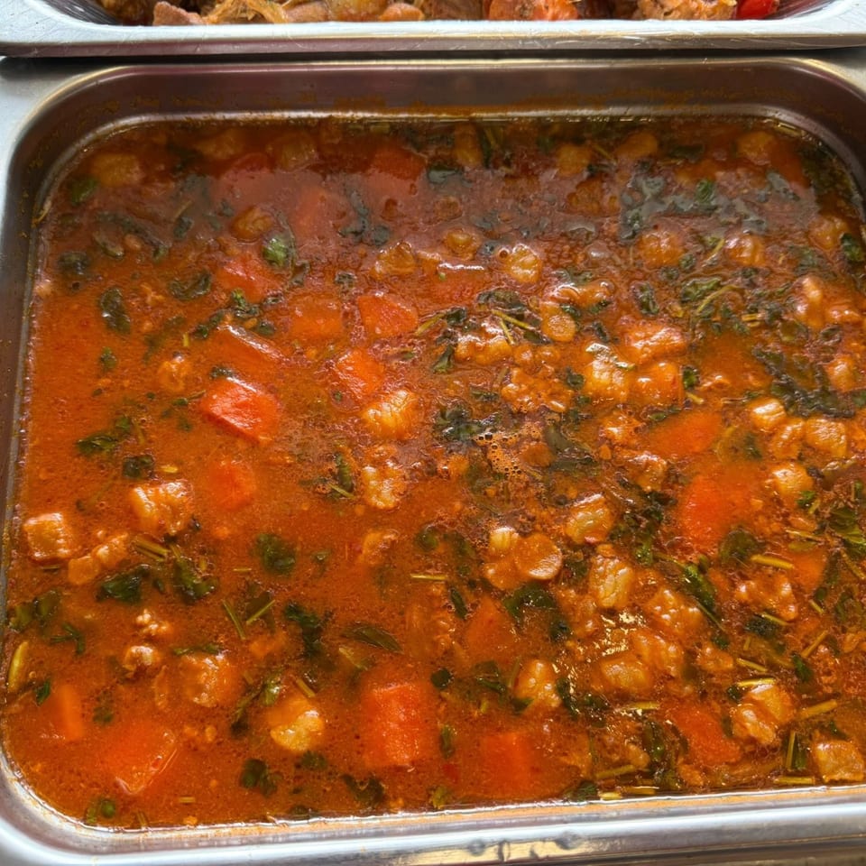 A close-up of a large metal tray filled with a rich, red Latin-American stew with vegetables.