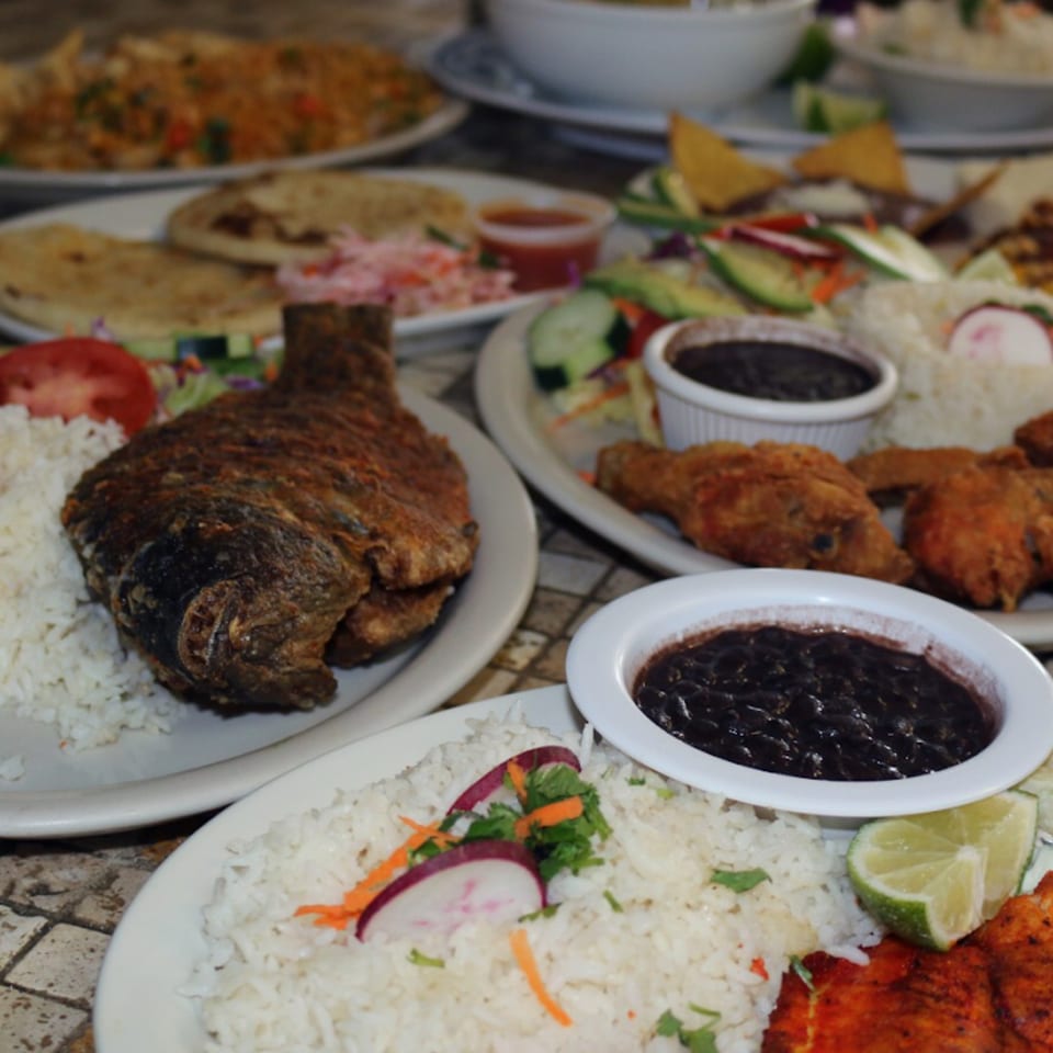 Plates of Latin American food, including fried fish, rice, black beans, fried chicken, and salads.