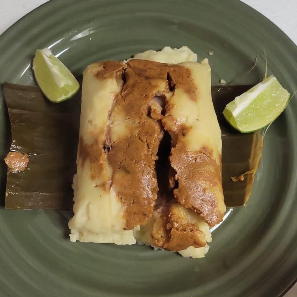 A Latin American tamale with a brown filling, served on a banana leaf with lime wedges.